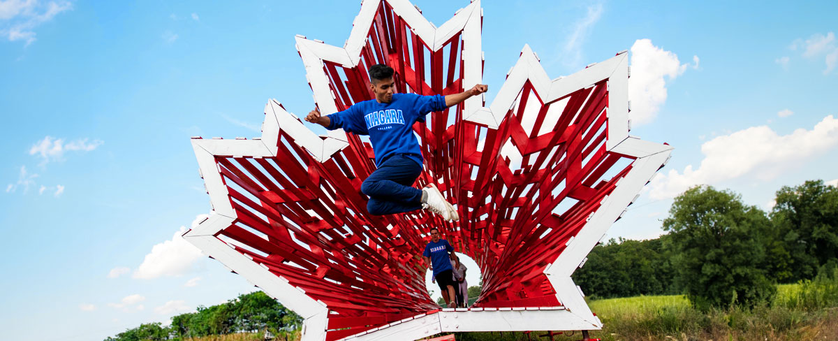 An international student wearing a Niagara College sweatshirt jumps in the air in front of a large red wooden maple leaf