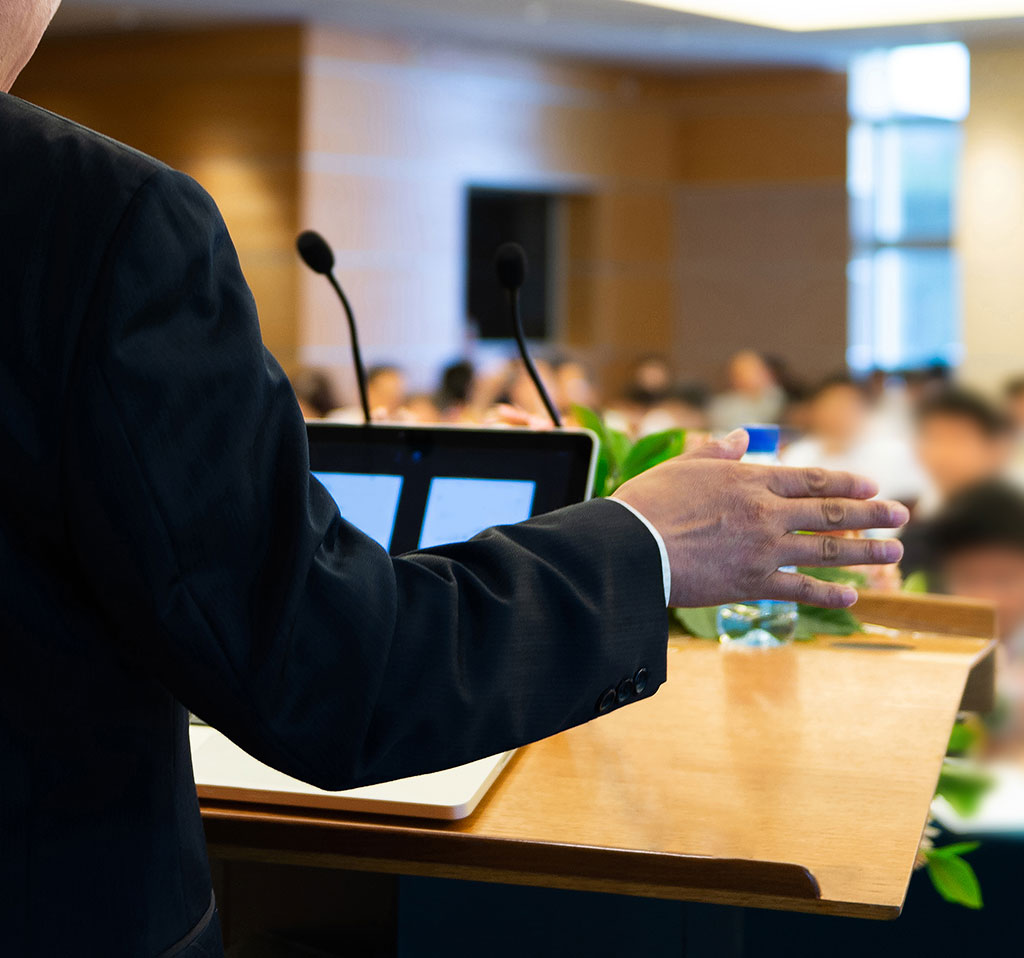 Man in a business suit standing at a podium gives a speech to a packed room